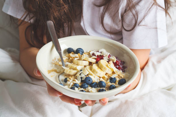 Overnight oatmeal with berries, banana, jam and coconut in white bowl in hands.